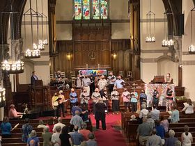 A congregation gathers in a church, in front of the altar and pipe organ and in several rows of pews.