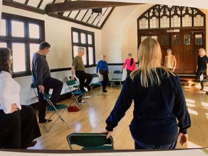 Yoga in the Headstart Room. In a brightly lit room with hardwood floors and exposed wood beams, eight adults in loose clothing stand on one leg while holding onto a chair.