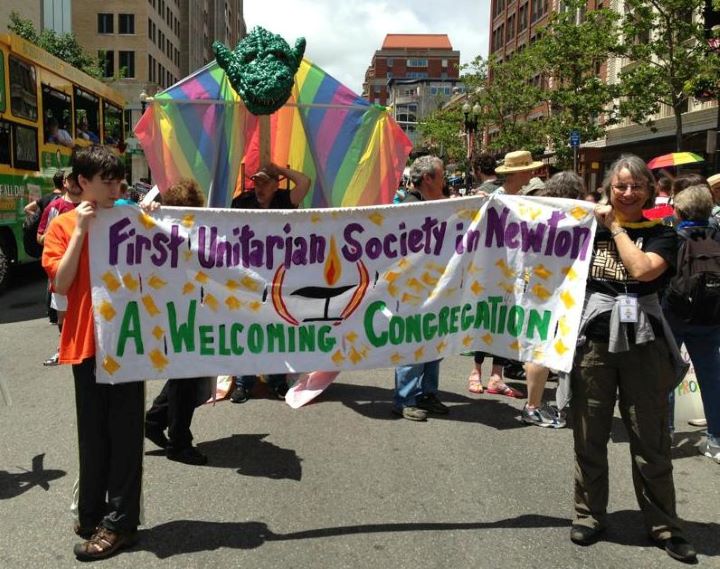 In the midst of a parade, a teenager and a woman with short gray hair hold up a banner that reads, 