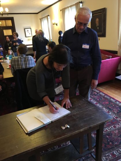 Signing the member book. In room with several people seated around a large table, a woman stands at a smaller table, signing a book, while a man looks on.