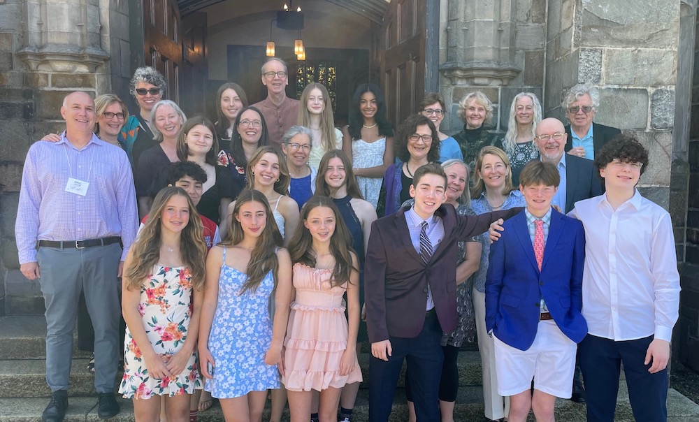 a group of youth and adults in colorful, dressy clothing stand, smiling, on the front steps of the church