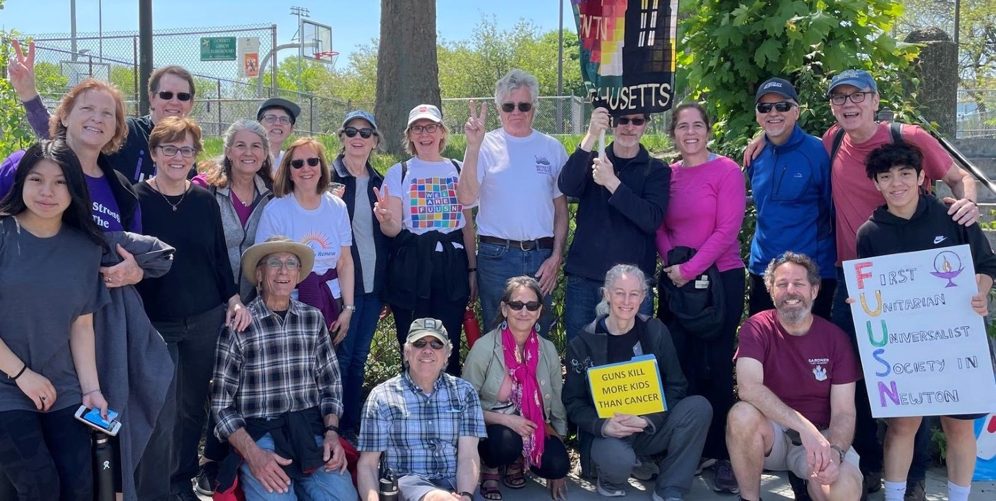 A large group of youth, adult, and elderly, many holding up a hand with the sign of peace, stand or kneel in a green space with trees, holding various signs and banners that read, 