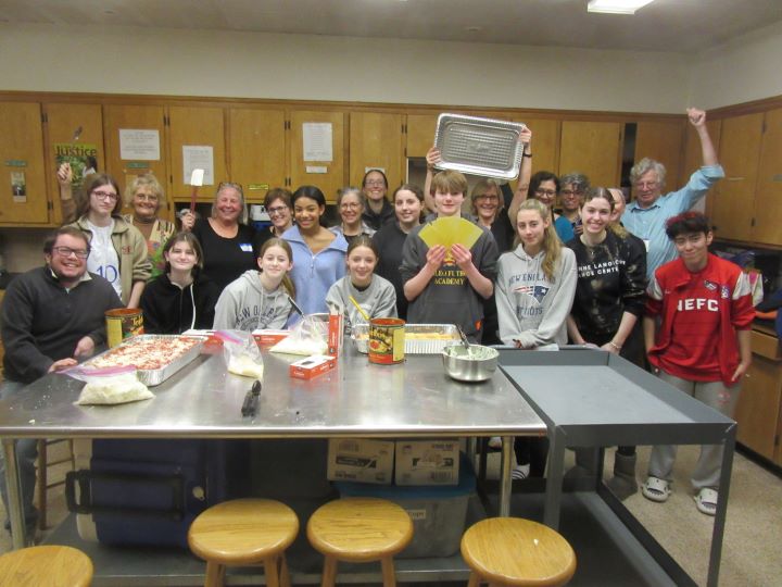 In a kitchen at a large stainless steel table, a group of about twenty adults and teenagers work together to cook lasagna for the homeless.