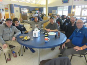 Six adults, including one woman with her arms outstretched in greeting, are seated around a round table in a cafeteria, with many other adults and children seated at tables behind them.