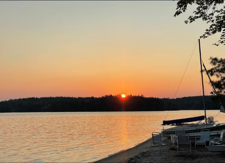 A vivid orange sunset as the sun slips behind wooded hills bordering Lake Winnepesauke, with a sailboat and lawn chairs on a sandy beach.