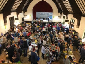 Shoppers in the Parish Hall browse through aisles of folding tables covered with yard sale items such as bags, umbrellas, kitchen appliances, and artificial Christmas trees.