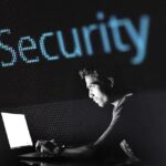 A man using a laptop in a dark room with the word "Security" suspended above him.