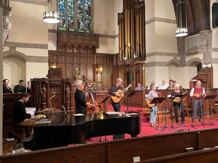 Five band members stand at the front of the church playing bass, guitar, and mandolin, accompanied by a pianist at a grand piano