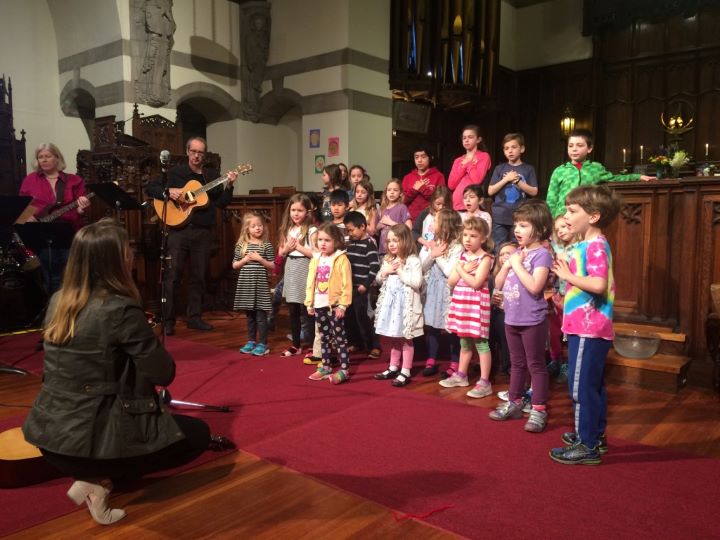 A choir composed entirely of children sings in the front of a church, directed by a kneeling woman with long brown hair wearing a black suit, and accompanied by a man with a guitar