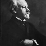 Black and white profile portrait of a man with white hair, mustache, and beard wearing a formal suit and tie with his arms crossed.