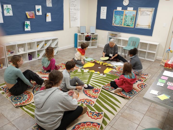 A gray haired woman with glasses wearing a striped shirt sits on the floor with a group of five children and two teenagers in a classroom.