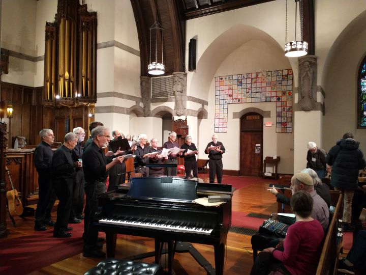 A group of eleven men dressed in back and holding binders stand at the front of the Sanctuary beside a grand piano