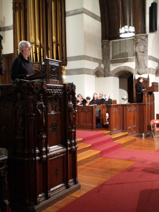 A gray-haired woman wearing glasses and a black long-sleeved shirt and a brown-haired woman dressed in black at a podium on the opposite side of the Sanctuary read together, with a choir seated behind them.