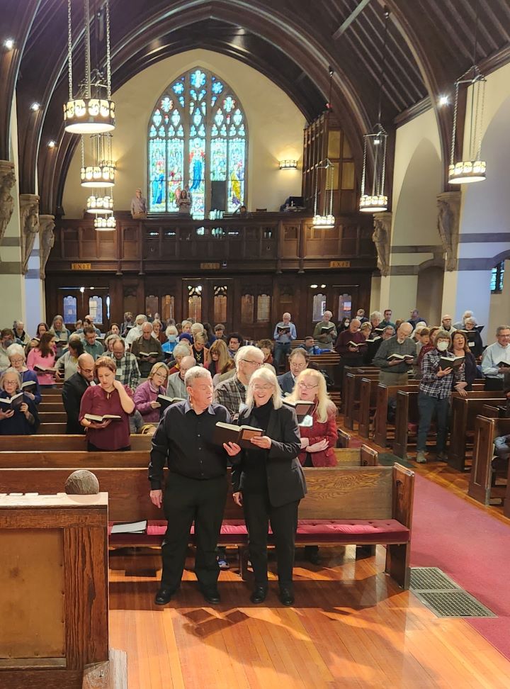 Music Director Anne Watson Born, a gray-haired woman with glasses wearing a black shirt and black suit, stands holding a hymnal with a congregant in the first pew of a church filled with singing congregants, including a balcony above the main sanctuary.