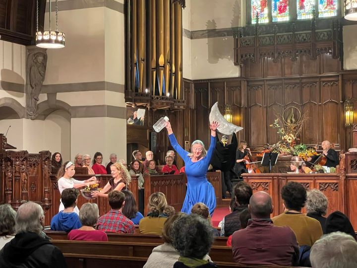 A troupe of dancers perform in the front of a church, including a woman with long gray hair wearing a long blue dress, her arms flung up with a water pitcher in one hand; a man in a black cape holding up a large, stuffed crescent moon; a woman with long, wavy blonde hair wearing a short-sleeved brown dress holds out a large glass bowl; another woman with shoulder-length brown hair wearing a white long-sleeved dress, white headband, and face mask, sprinkles cloth flowers into the outstretched bowl