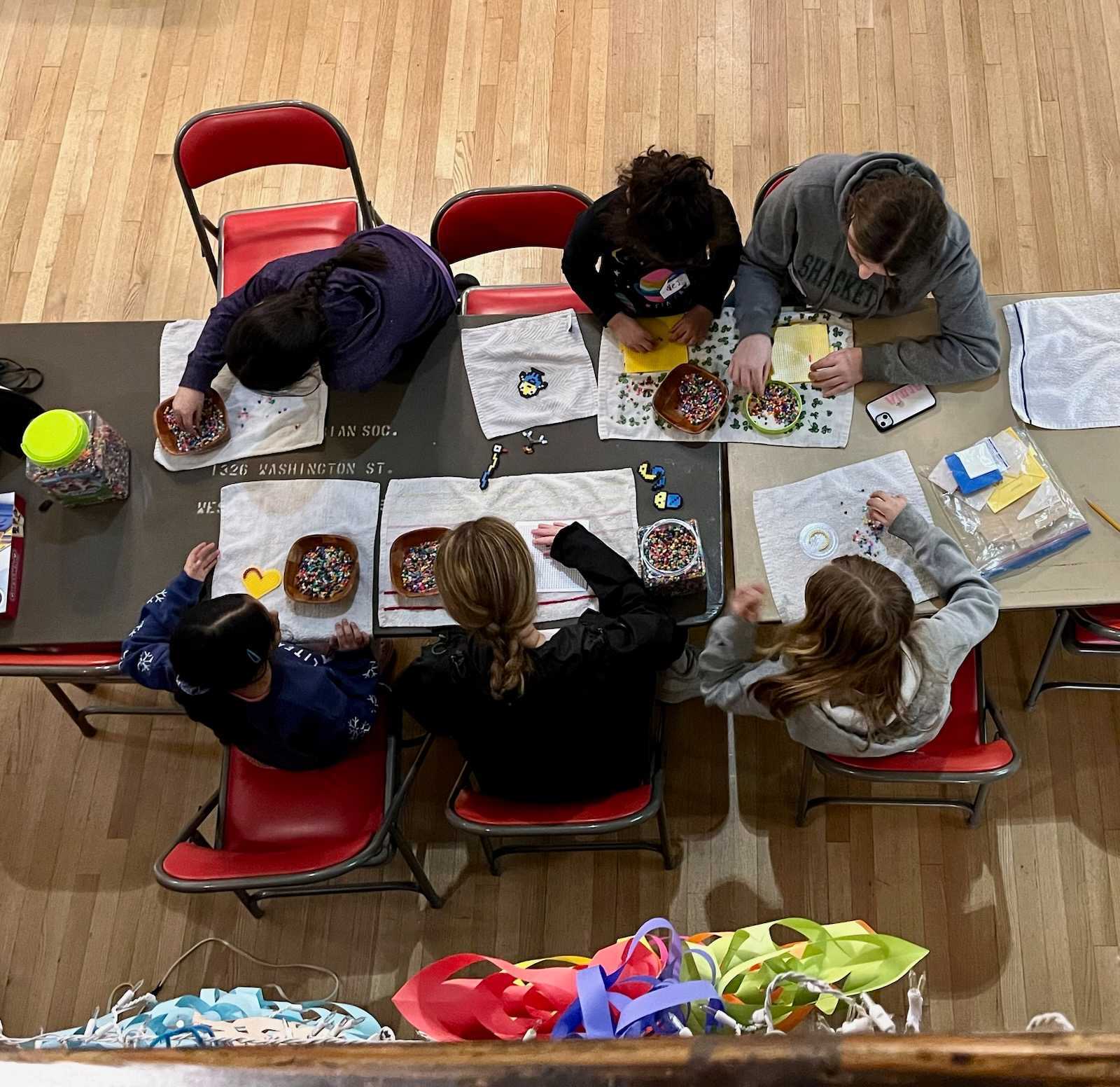 Six children sit at a table creating crafts with beads.