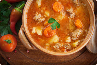 A brown ceramic crock with two handles contains soup with chunks of meat, carrots, potato, and a single leaf of potato. On the wooden table next to the crock are a whole tomato, whole chile pepper, parsley leaves, pomegranate seeds, and a carved wooden spoon. 