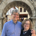 A gray-haired man with glasses in a blue button-up shirt and a woman with chin-length blonde hair and glasses in a black sleeveless top with large buttons down the side stand outside under a stone archway.