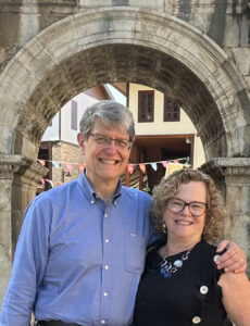 A gray-haired man with glasses in a blue button-up shirt and a woman with chin-length blonde hair and glasses in a black sleeveless top with large buttons down the side stand outside under a stone archway.