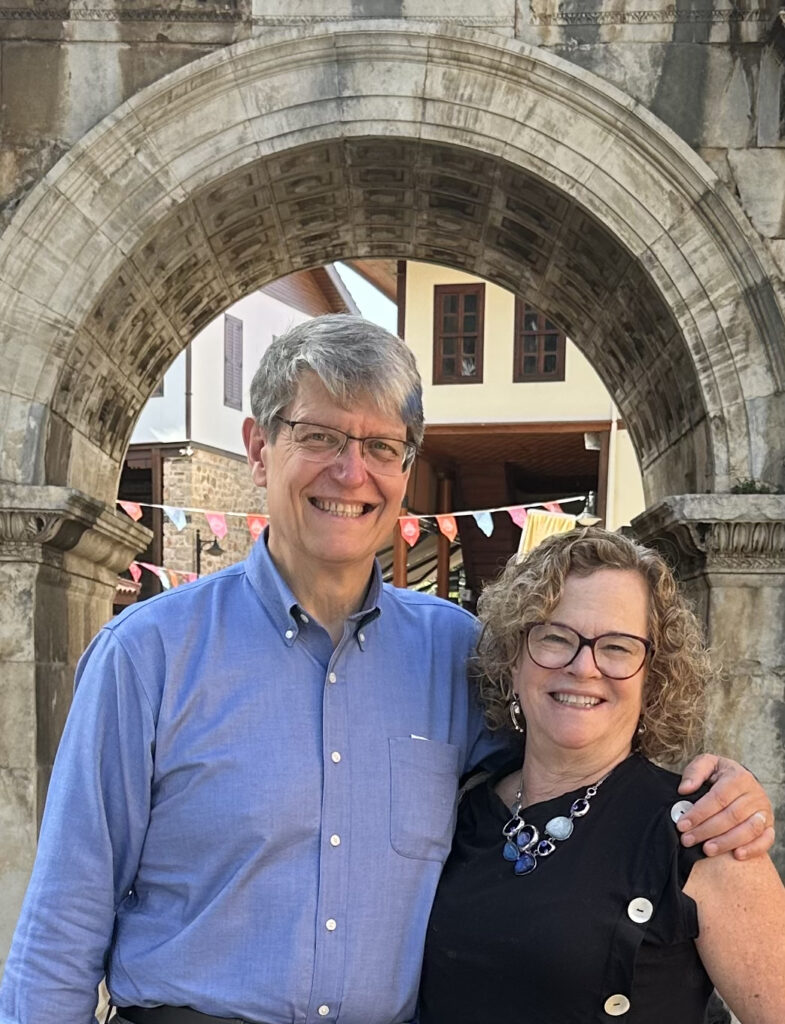 A gray-haired man with glasses in a blue button-up shirt and a woman with chin-length blonde hair and glasses in a black sleeveless top with large buttons down the side stand outside under a stone archway.