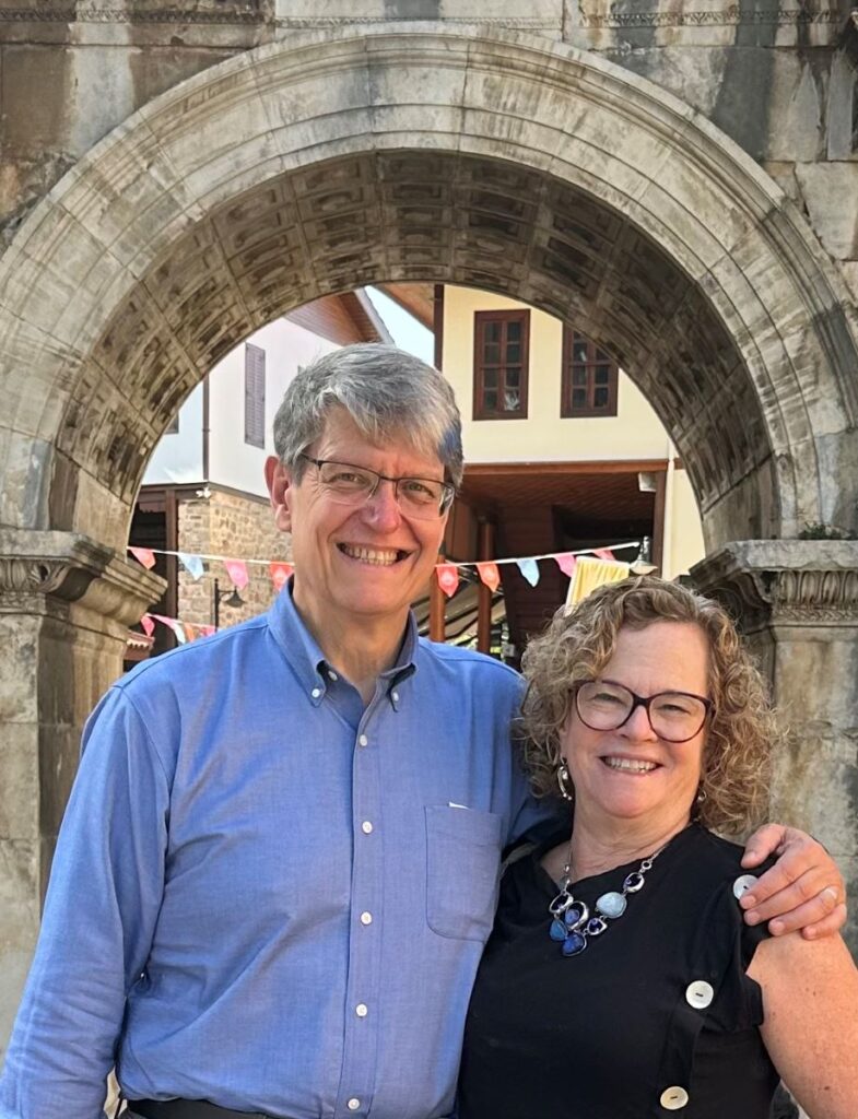 A gray-haired man with glasses in a blue button-up shirt and a woman with chin-length blonde hair and glasses in a black sleeveless top with large buttons down the side stand outside under a stone archway.