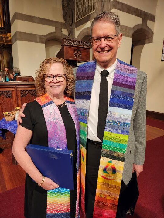 A woman with blonde hair and glasses on the left, wearing a black dress and rainbow ministerial stole, is embraced by a gray-haired man on the right wearing glasses, a gray suit jacket, white shirt, black tie, black pants, and rainbow ministerial stole.