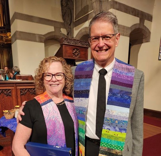 A woman with blonde hair and glasses on the left, wearing a black dress and rainbow ministerial stole, is embraced by a gray-haired man on the right wearing glasses, a gray suit jacket, white shirt, black tie, black pants, and rainbow ministerial stole.