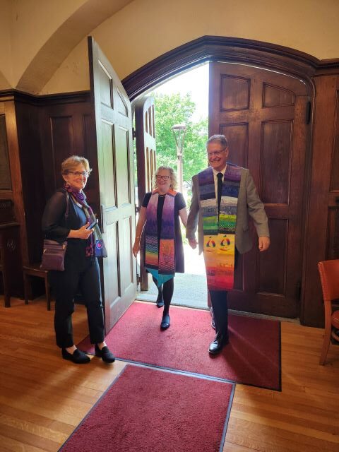 A blond woman and a gray-haired man, both wearing ministerial stoles, hold hands as the enter a church's narthex door, being held by a short-haired woman with glasses