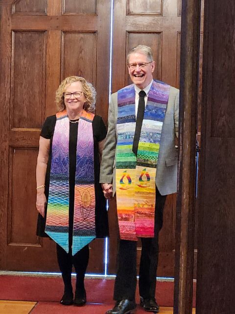 A woman with blonde hair and glasses on the left, wearing a black dress and rainbow ministerial stole holds hands with a gray-haired man on the right wearing glasses, a gray suit jacket, white shirt, black tie, black pants, and rainbow ministerial stole as they wait in the narthex to enter the sanctuary