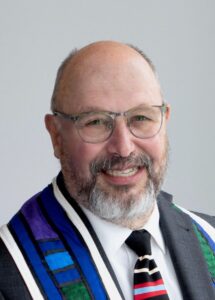 A smiling man with glasses and a gray beard wearing a suit, white shirt, ministerial stole, and a colorfully striped tie.