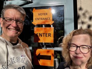 A gray haired man with glasses wearing a gray hoodie and a blonde woman with glasses wearing a black shirt stand in front of a glass door with yellow signs reading, "Voter Entrance" and "Enter" with an arrow pointing left.