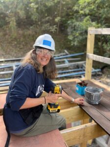 A woman with long wavy brown hair wearing glasses, a white hard hat, clear safety goggles, a navy blue sweatshirt that reads, "UMASS," and khaki pants holds a power drill and sits on a partially constructed balcony with her legs dangling between the wooden beams