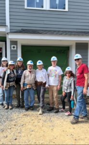 Eight adults in hard hats, some holding tools, stand in front of a partially constructed home.