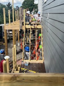 Two men wearing white hardhats, one wearing a gray short-sleeved athletic shirt and the other wearing a white T-shirt with a colorful graphic reading, "WE ARE FUUSN" sit on an unfinished balcony, their legs dangling through the beams behind an unfinished staircase being held by a third man in a blue hardhat and grey T-shirt.