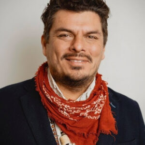 Headshot of a smiling man with brown hair, mustache, and beard wearing a dark blue woven jacket, white shirt, and multiple beaded necklaces covered by a red bandana.