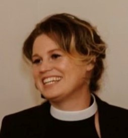 A smiling woman with short blonde hair wearing a black jacket, black sleeveless shirt, and white clerical collar