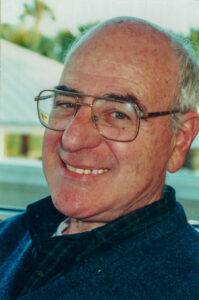 Headshot photo of a smiling man with gray hair and black eyebrows, wearing gold-framed glasses, a blue plaid shirt, and a blue knit sweater.